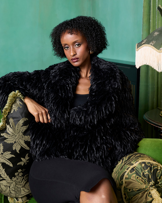 Woman wearing a black fur coat sitting on a decorative chair with a green curtain background